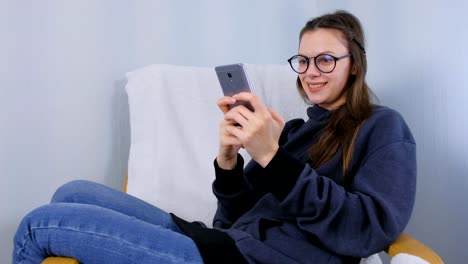 Junge-brünette-Frau-in-Brille-tippt-eine-Nachricht-auf-einem-Handy-sitzen-im-Sessel.