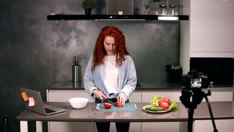 Mujer-joven-con-ropa-casual-dirige-un-blog-culinario-y-graba-vídeo-en-cocina-gris.-Video-shooting-del-blog---cabeza-roja-cortando-tomates-en-un-tablero.-Marketing-moderno,-redes-sociales