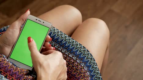 Close-up-woman-in-a-skirt-using-smartphone-with-green-screen-while-sitting-on-the-couch-in-the-livinng-room.-Woman-hands-scrolling,-typing,-tapping-on-touchscreen.-chroma-key.-top-view.-4K-UHD.-Closeup.