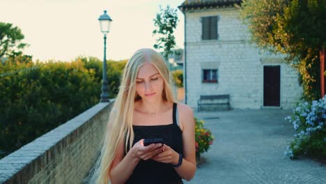 Charming-young-woman-using-smartphone-while-walking-outside-in-summer