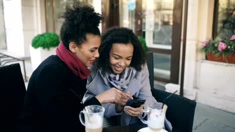 Dos-atractivas-amigas-de-raza-mixta-compartiendo-juntas-usando-smartphone-en-el-café-callejero-al-aire-libre