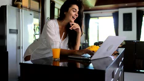 Chica-uso-portátil-en-cocina-chateando-en-línea-joven-bebida-jugo-estudio-casa-moderna-Interior