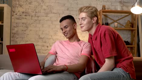 Two-multinational-homosexual-friends-sit-on-the-couch,-use-a-laptop,-show-a-gesture-of-goodbye,-air-kiss-in-a-video-chat.-Home-cosiness,-family,-internet-concept.-60-fps