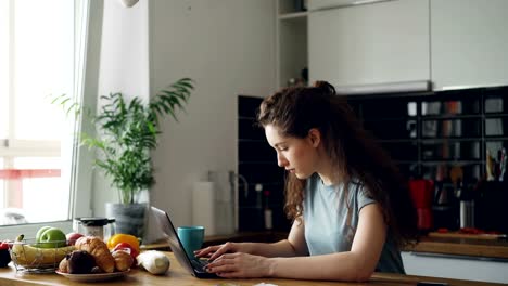 Young-concentrated-cheerful-woman-working-in-kitchen-typing-laptop-computer-during-breakfast-in-the-morning