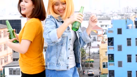 Mujer-asiática-joven-pareja-de-lesbianas-bailando-y-tintineantes-botellas-de-cerveza-fiesta-en-la-azotea.