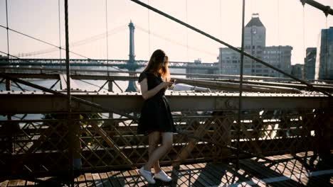 Beautiful-Caucasian-woman-stands-using-smartphone-messenger-app-at-Brooklyn-Bridge,-New-York-amazing-skyline-panorama-4K