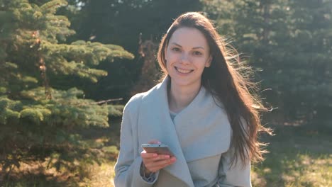 Joven-hermosa-mujer-morena-se-sienta-en-la-hierba-en-el-parque-de-la-primavera-y-tipos-de-un-mensaje-en-el-teléfono-móvil,-mira-a-cámara-y-sonriendo.