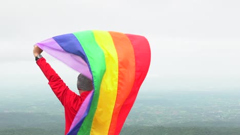 hombre-levantar-bandera-LGBTI-de-color-arco-iris-ondeando-en-duro-viento-en-el-mirador-de-alta-montaña