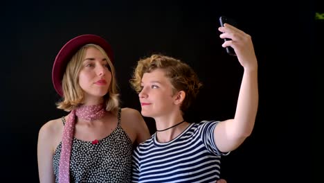 Two-charming-young-blonde-women-hugging-and-taking-selfie,-standing-isolated-on-black-background