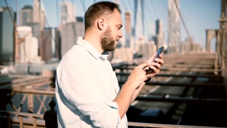 Happy-European-businessman-using-smartphone-mobile-office-app-at-Brooklyn-Bridge,-then-turning-away-from-camera-4K