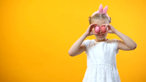 Happy-little-girl-holding-colored-Easter-eggs-in-front-eyes-and-smiling-camera