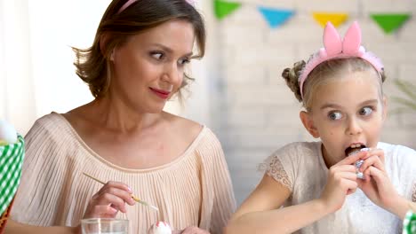 Schelmischen-Mädchen-Ostern-Schokolade-zu-essen,-während-Mutter-malen-Eier,-Kindheit