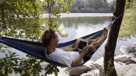 Junger-Mann-mit-digital-Tablette-auf-Hängematte,-liegend-zwischen-den-Bäumen-am-Fluss-im-Sommer