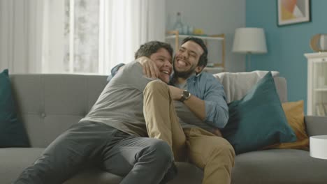 Sweet-Male-Gay-Couple-Full-Around-on-a-Sofa-at-Home.-Boyfriend-Runs-and-Jumps-into-Hands-of-His-Partner.-They-Hug-and-Look-at-Each-Other.-They-are-Happy-and-Laughing.-Room-Has-Modern-Interior.