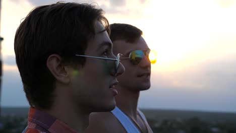 Profile-of-young-male-couple-in-sunglasses-standing-on-the-edge-of-rooftop-and-talking.-Handsome-gay-boys-relaxing-on-roof-of-high-rise-building-and-enjoying-beautiful-cityscape.-Close-up-Slow-motion