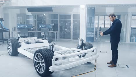 Engineer-with-Glasses-and-Beard-Scans-Electric-Car-Chassis-Prototype-with-Wheels,-Batteries-and-Engine-with-an-Augmented-Reality-Software-on-a-Tablet-Computer-in-a-High-Tech-Development-Laboratory.