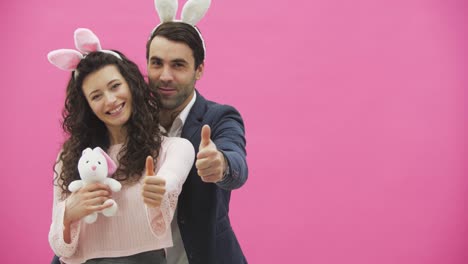 Young-couple-standing-on-a-pink-background.-During-this-show-a-gesture-class-smiling-and-holding-a-soft-toy-in-the-hands-of-a-bunny.-Happy-family-is-preparing-for-Easter,-with-the-ears-of-a-pink-rabbit-on-its-head.