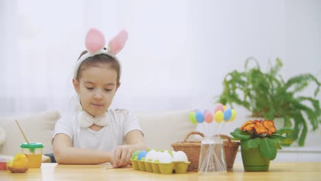 Little-cute-girl-is-having-fun-holding-a-paint-brush-in-her-left-hand.-Girl-with-a-beauty-spots-at-her-face-watching-at-nude-paint-brushes,-sitting-at-the-wooden-table-with-Easter-decorations.-Girl-shows-her-fingers-up.