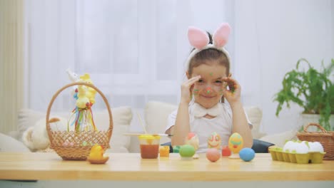 La-niña-linda-con-orejas-de-conejo-está-ocultando-su-hermosa-cara.-Suddelry-muestra-su-coloreado-en-colores-brillantes-cara-y-sonriendo-ampliamente,-mostrando-sus-dientes.