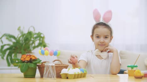 Kleine-niedliche-und-entzückende-Mädchen-lächelt-aufrichtig.-Sie-nimmt-ein-Osterei-aus-dem-Korb-und-schaut-es-an-und-zeigt-dann-darauf.-Konzept-Osterferien.