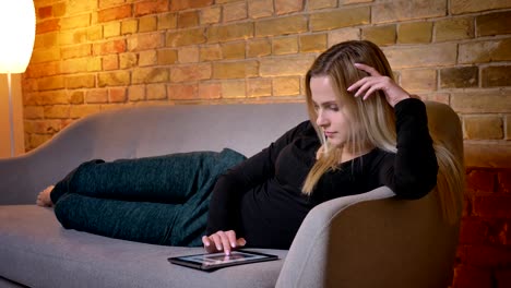 Nahaufnahme-Porträt-der-jungen-hübschen-Hausfrau-mit-dem-Tablet-und-Liegen-auf-dem-Sofa-lehne-zurück-in-Innenräumen-in-gemütlichem-Haus