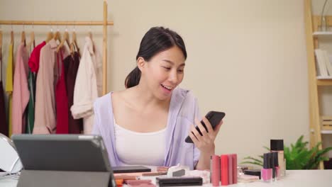 Beauty-Blogger-Frage-und-Antwort-per-Telefon-sitzen-vor-Tablet-für-die-Aufnahme-von-Video.-Glücklich-schöne-junge-asiatische-Frau-verwenden-Kosmetik-Rezension-Make-up-Tutorial-übertragen-Live-Video-an-soziale-Netzwerk.