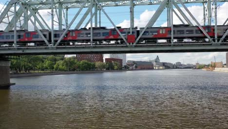 Luftaufnahme-der-Eisenbahnbrücke,-mit-einem-fahrenden-Zug-auf-ihr,-über-den-Fluss,-der-durch-eine-Großstadt-fließt