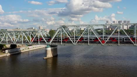 Vista-aérea-del-puente-ferroviario,-con-un-tren-en-movimiento-en-él,-a-través-del-río-que-fluye-a-través-de-una-ciudad-importante