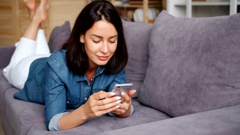 Smiling-young-lady-using-smartphone-holding-gadget-touching-screen-at-home