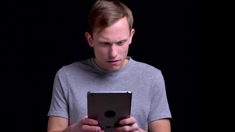 Closeup-portrait-of-young-handsome-caucasian-man-typing-on-the-tablet-in-front-of-the-camera-with-background-isolated-on-black