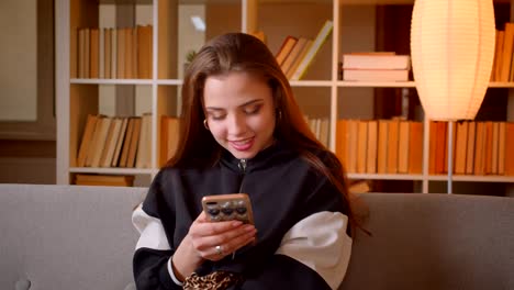 Portrait-of-young-teenage-girl-watches-into-cellphone-sitting-on-sofa-on-bookshelves-background-at-home.