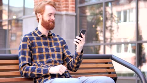 Rotschopf-Bart-junger-Mann-feiert-Sieg-auf-dem-Smartphone,-sitzen-im-Freien-auf-Bank