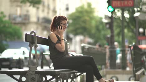 Cute-smiling-redhead-girl-with-glasses,-freckles,-piercings-and-red-hair-having-phone-conversation-on-her-smartphone-sitting-on-street-bench,-during-sunny-summer-in-Paris.-4K-UHD.-Trendy.