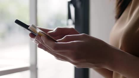 Close-up-hands-of-a-beautiful-Asian-caucasian-woman-using-smartphone-shopping-online-and-use-the-credit-card-payment-while-standing-beside-the-window-at-the-office-room.