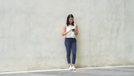 Disparo-de-cuerpo-completo-de-feliz-joven-hermosa-mujer-inclinada-en-la-pared-mientras-usa-el-teléfono-al-aire-libre