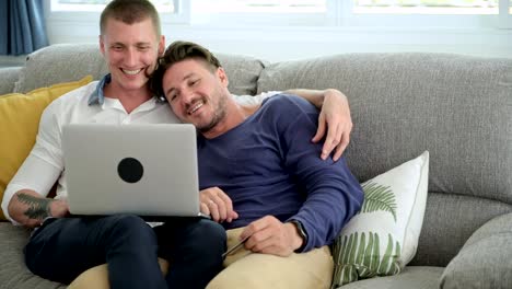 Gay-couple-relaxing-on-couch-using-laptop-computer.-Happy-shopping-and-kissing.