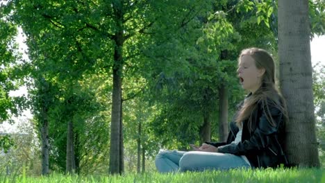 Young-beautiful-girl-in-street-clothes-in-a-park.-Holding-a-smartphone-in-his-hands,-sitting-on-the-grass-under-a-tree.-Reads-boring-message-and-photos,-Yawns-heavily.-Not-enough-sleep,-wants-to-sleep