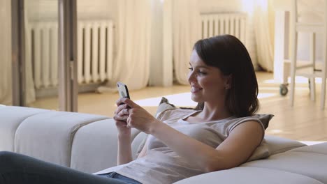 Smiling-relaxed-young-woman-holding-smartphone-texting-at-home