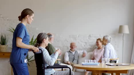 Weibliche-Krankenschwester-schieben-Rollstuhl-mit-behinderten-älteren-Frau.-Senior-Patient-nimmt-Pillen-und-Glas-Wasser-auf-dem-Tisch-und-mit-Freunden-sprechen-auf-dem-Sofa-im-Gemeinschaftsraum-des-Pflegeheims