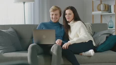 Cute-Young-Couple-Use-Laptop-Computer,-Have-Fun,-Laughs,-while-Sitting-on-the-Couch-in-the-Cozy-Apartment.-Couple-Shopping-on-Internet,-Using-Social-Media,-Watching-Videos-and-Streaming-Content