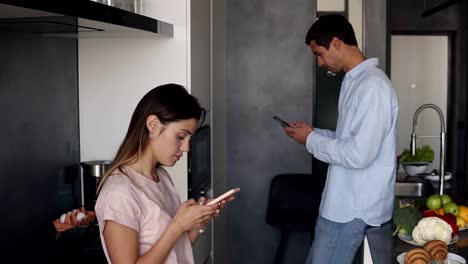 Pareja-joven-mirando-algo-en-la-pantalla-de-sus-teléfonos-inteligentes-en-la-cocina-con-diseño-loft-moderno.-Leer-noticias-de-la-mañana-o-consultar-las-redes-sociales