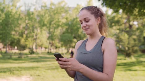 Caucasian-fit-young-woman-smiling-while-texting-on-mobile-phone-at-the-park-in-the-morning-on-a-sunny-day