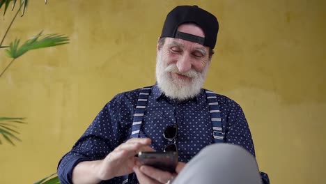 Front-view-of-handsome-high-spirited-70-aged-beard-man-in-stylish-cap-which-sitting-near-yellow-wall-and-using-his-mobile