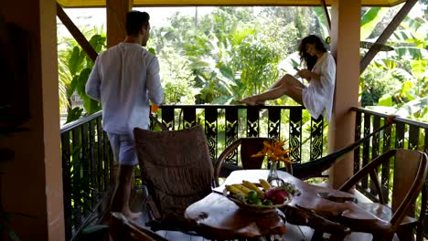 Hombre-hacer-jugo-celular-inteligente-de-uso-de-joven-sentado-en-el-balcón,-feliz-pareja-hablando-en-terraza-de-verano