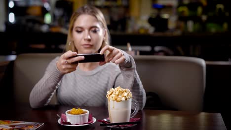 fröhliche-junge-Frau-mit-blonden-Haaren-ist-ihr-Kuchen-und-Kaffee-per-Telefon,-im-gemütlichen-Kaffeehaus-sitzen-fotografieren