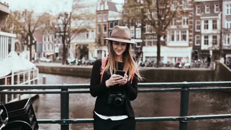 Hübsche-Frau-mit-Smartphone-auf-einer-Brücke.-4-K.-junge-Dame-im-stylischen-Hut-mit-langen-Haaren-und-Kamera-Typen-eine-Nachricht