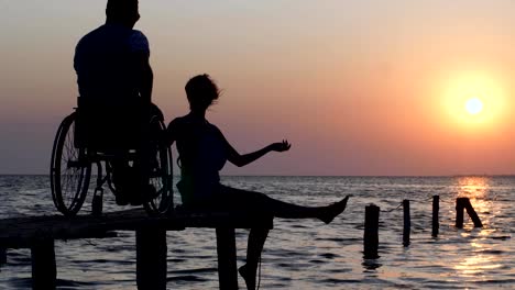 lachende-Mädchen-sitzen-auf-Pier-und-im-Gespräch-mit-Mann-im-Rollstuhl-Kulisse-von-Meer-und-Sonnenuntergang-deaktiviert