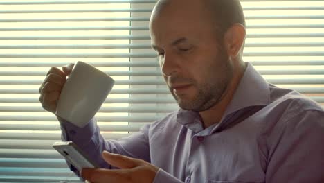 Tired-Businessman-standing-with-smartphone-by-window-in-the-office.-Close-up,-serious-man-sitting-by-out--window-and-uses-a-smartphone.-Thoughtful,--problem,-social-network.-Boss-drinks-coffee,-tea.