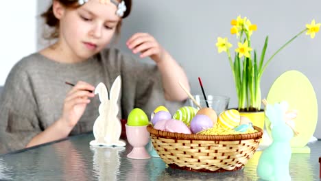 La-chica-hermosa-pinta-la-figura-del-conejito-de-Pascua.-Hay-pinturas-sobre-la-mesa,-una-cesta-con-huevos-de-Pascua-y-un-ramo-de-narcisos