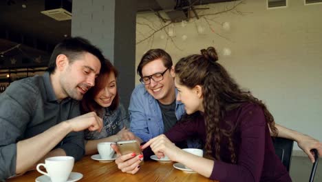 Los-jóvenes-están-viendo-smartphone-pantalla-discutiendo-fotos-y-riendo-durante-el-almuerzo-en-la-cafetería.-Amistad,-tecnología-moderna-y-el-concepto-de-estilo-de-vida-moderno.